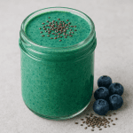 Grün-blauer Smoothie in einem Glas, getoppt mit Chiasamen; daneben liegen frische Blaubeeren und Chiasamen auf einem hellen Untergrund