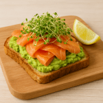 Lachs-Avocado-Toast auf Holzbrett mit cremiger Avocado, geräuchertem Lachs, frischen Brokkoli-Sprossen und Zitronenspalte